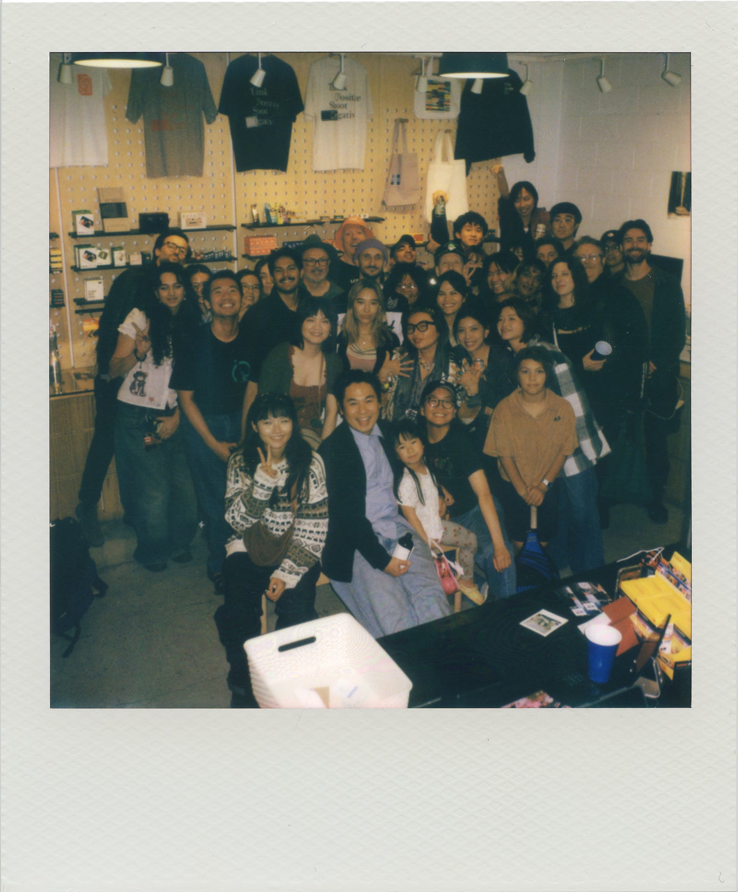 A group photos of turnout together at the previous After Hours event, captured in a Polaroid-style photo with a white border. Several black t-shirts are hanging on display in the background.