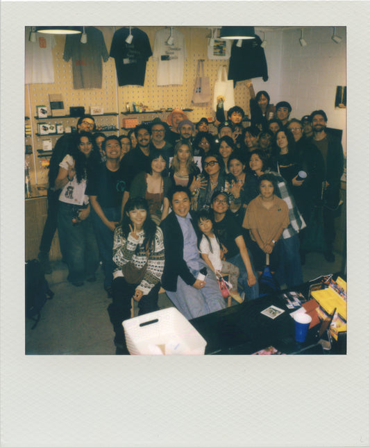 A group photos of turnout together at the previous After Hours event, captured in a Polaroid-style photo with a white border. Several black t-shirts are hanging on display in the background.